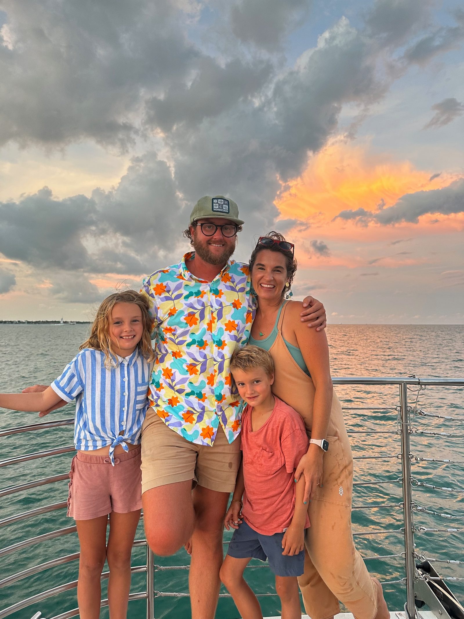 A family of four posing on the bow of the boat at sunset after the trip, with a pink and gray sky behind them