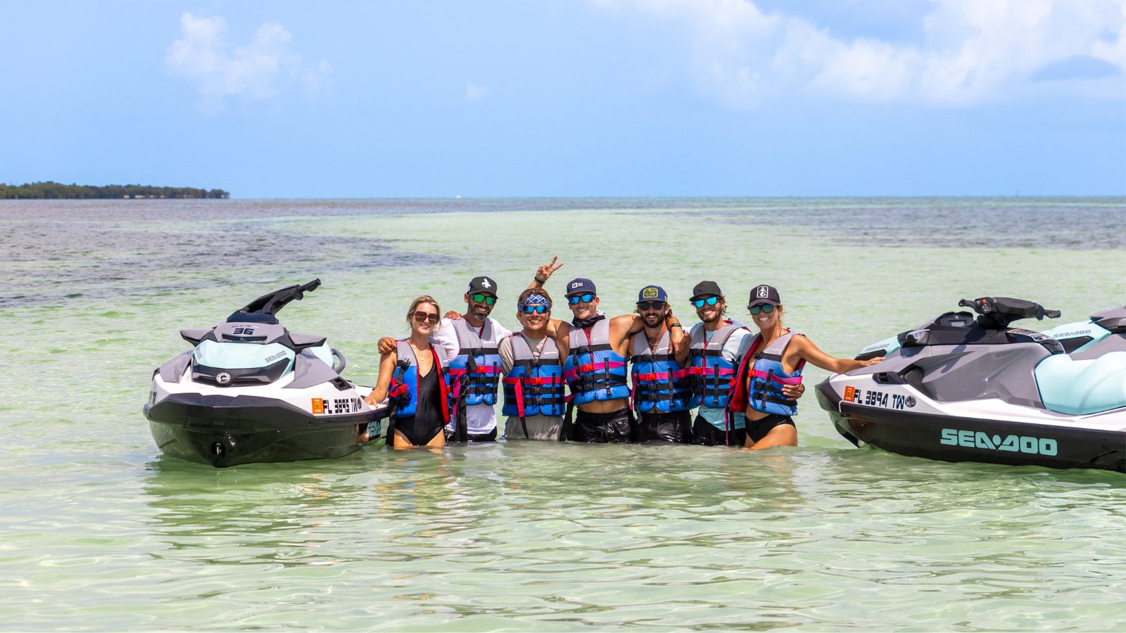 A group of riders in life jackets standing waist-deep on a sandbar between two parked jet skis, smiling for a photo