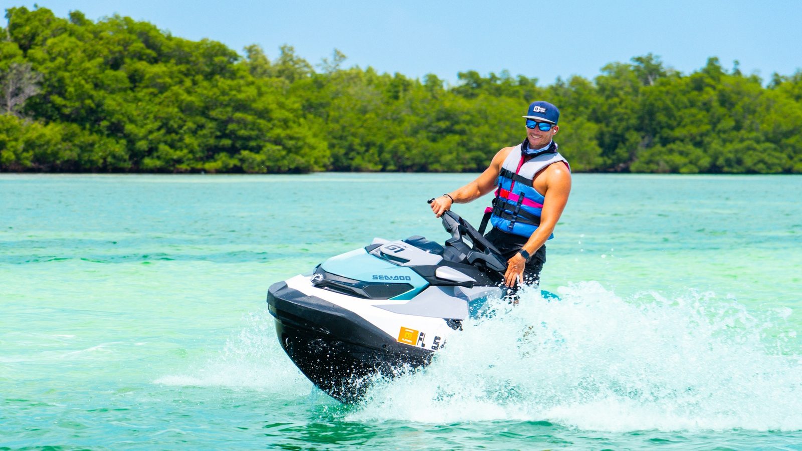 A rider in a life jacket carving a turn on a Sea-Doo with spray flying and a mangrove shoreline behind
