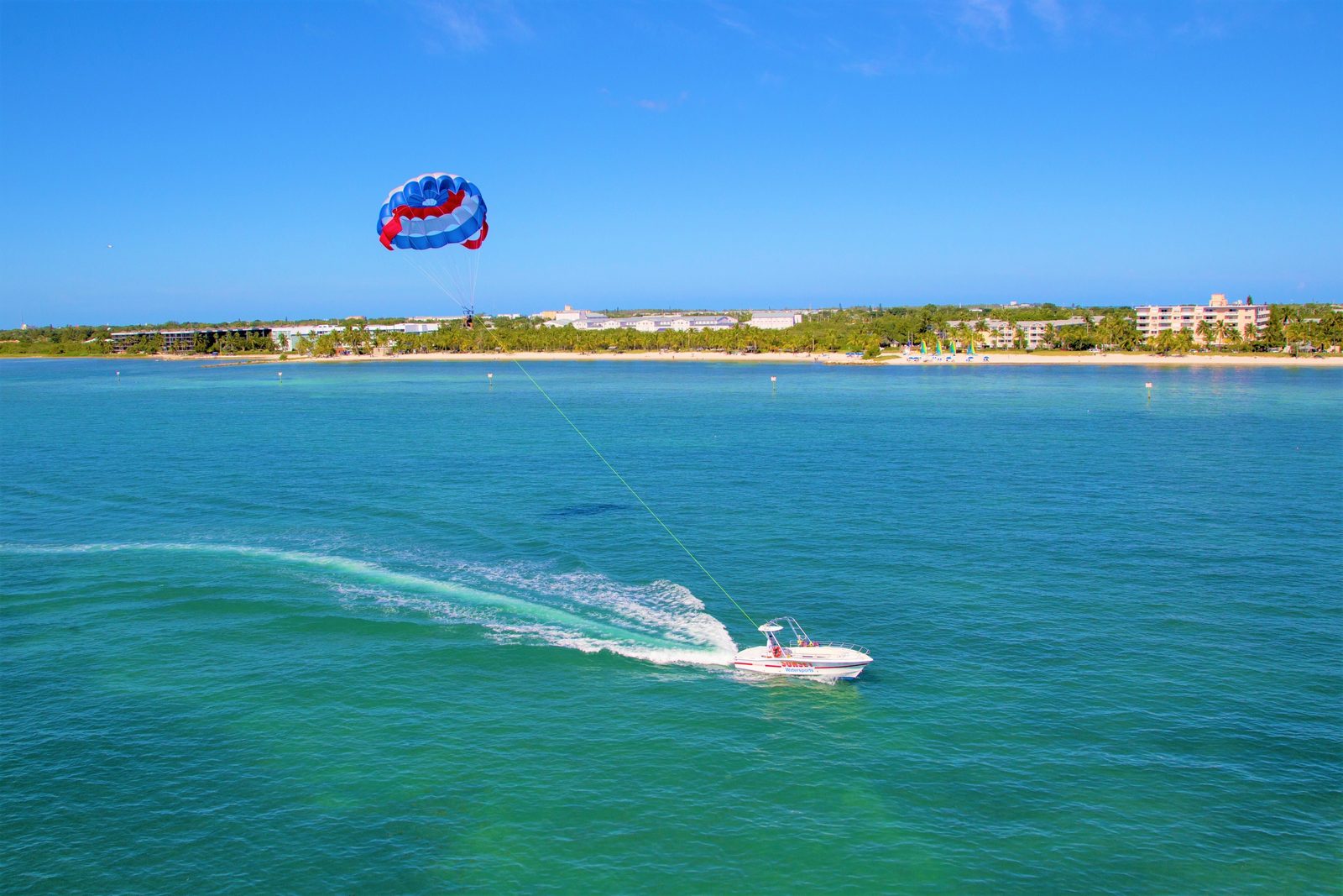 Aerial view of the parasail boat towing a red, white and blue canopy along the Key West shoreline with hotels and palms behind