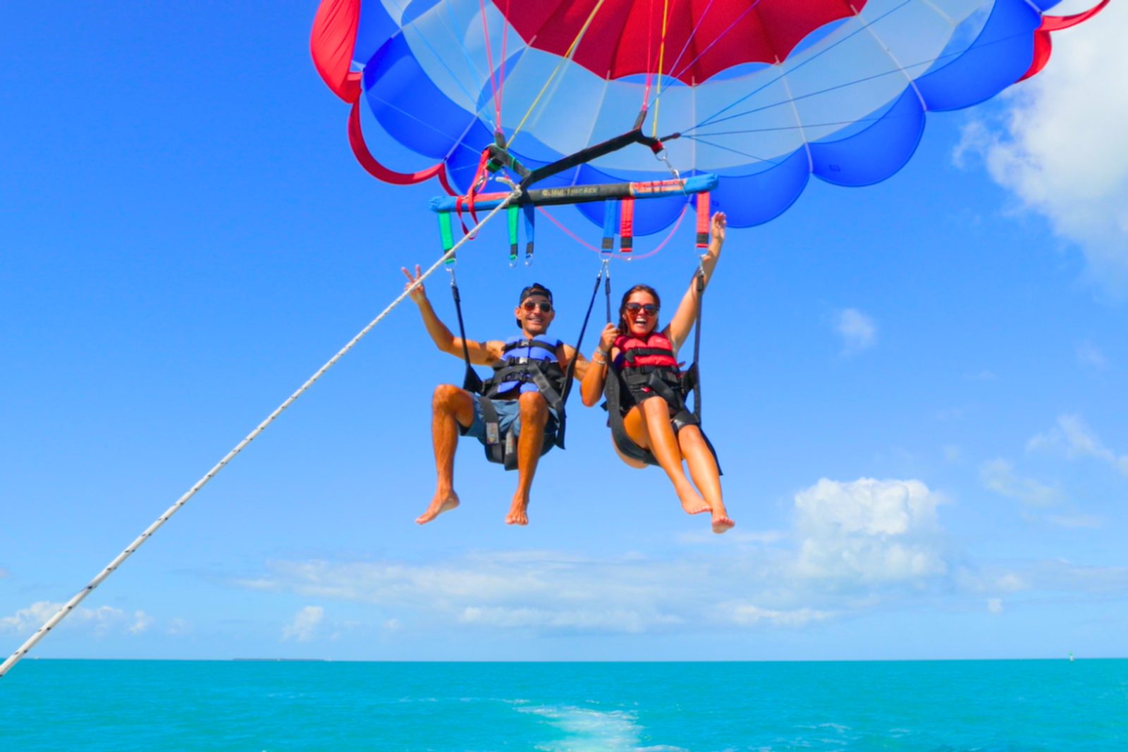 A couple in harnesses under the parasail canopy with their feet dangling over flat turquoise water