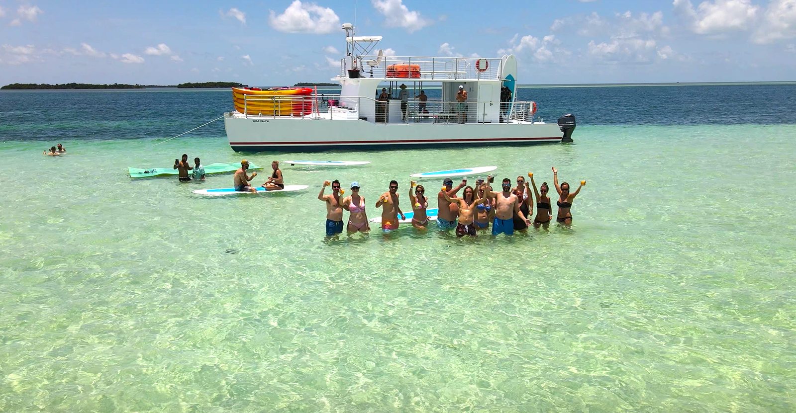 A group of guests standing in shallow clear water in front of the catamaran at the sandbar, arms raised for a photo