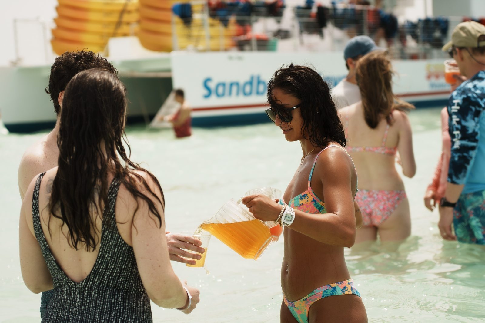 Two guests in swimsuits holding drinks in the shallows beside the catamaran, with other guests in the water behind