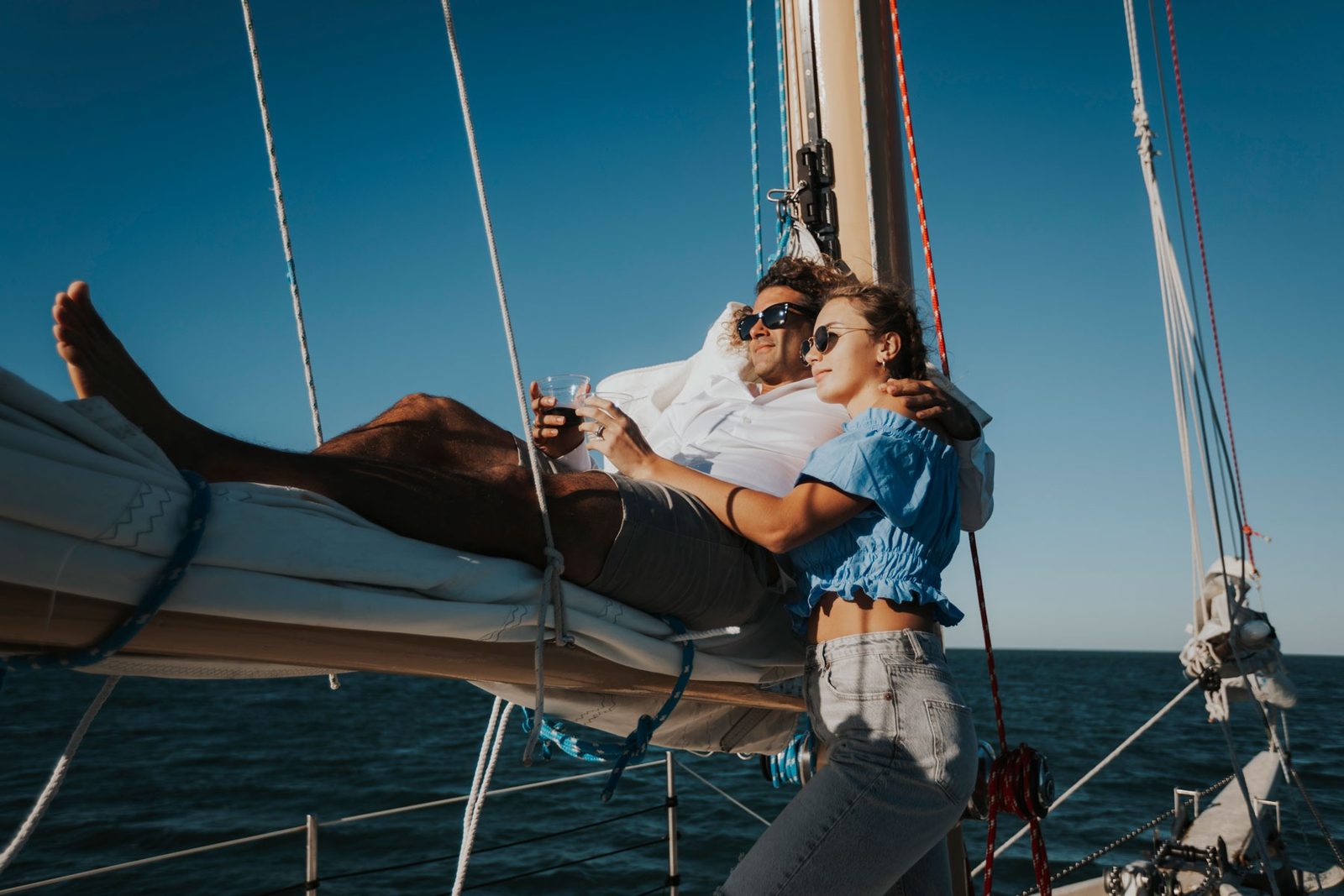 A couple leaning back on the bowsprit of a schooner with wine glasses, sails and rigging above them against a blue sky