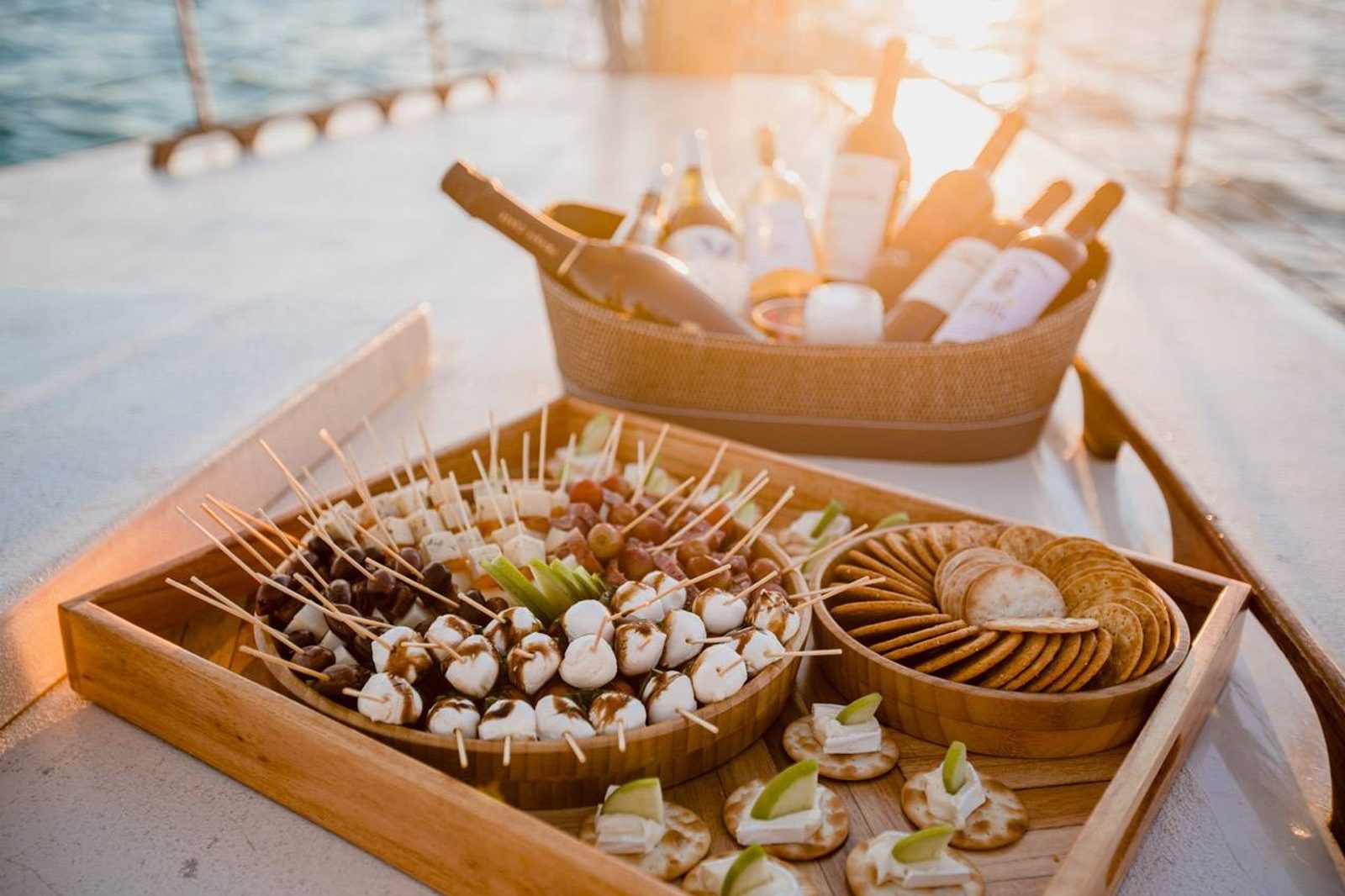 Wooden trays of charcuterie, cheese, caprese skewers and crackers laid out on deck beside a bucket of wine bottles in golden light