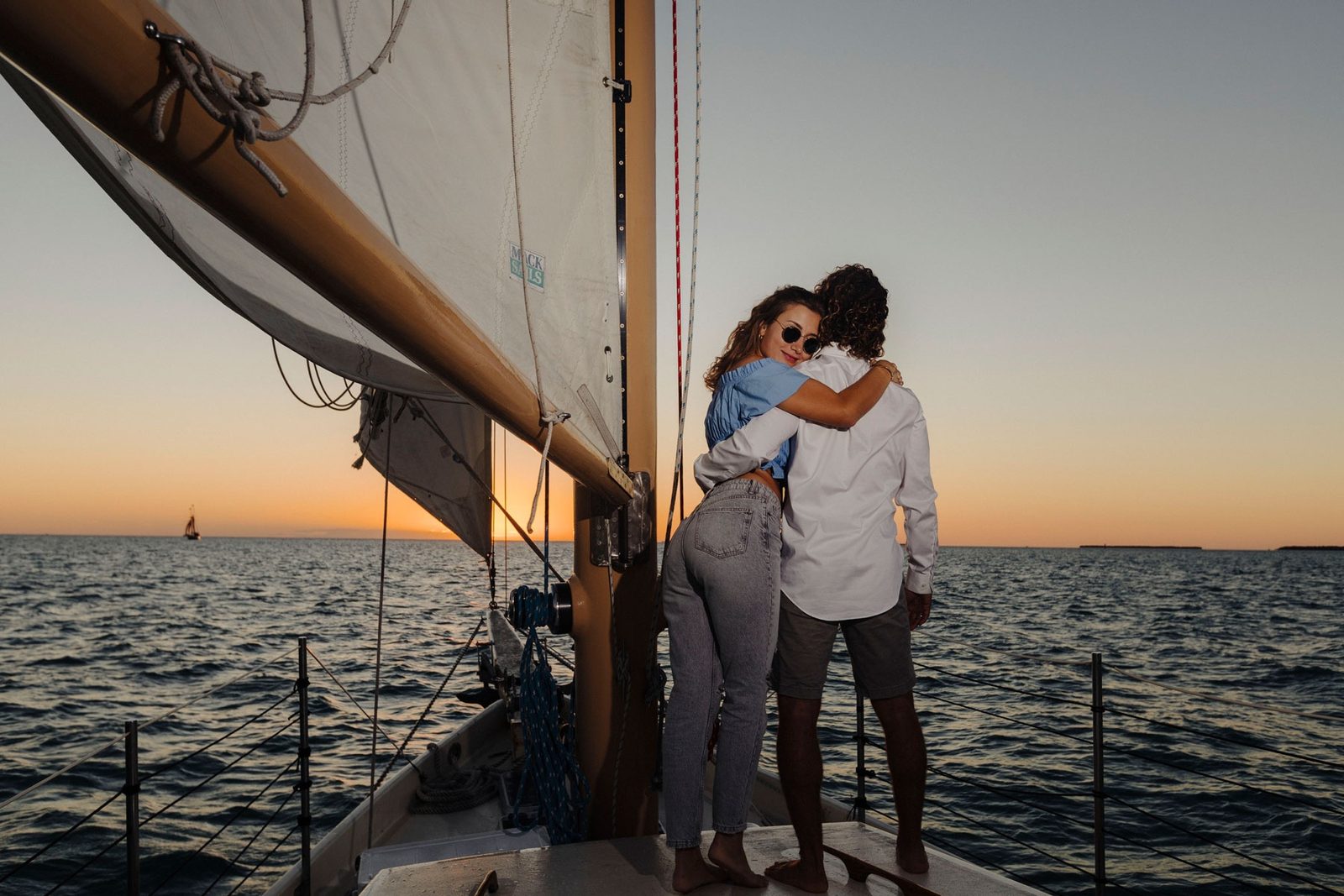 A couple embracing on the foredeck of a schooner at dusk with the sails up and the water glowing orange